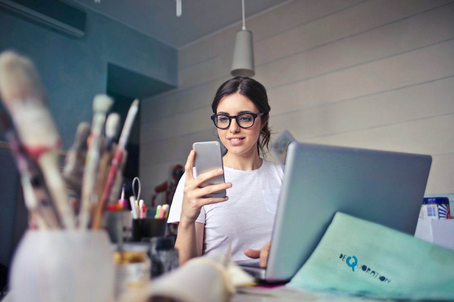 A person working at a laptop in a small studio, holding a phone, with brushes in the foreground