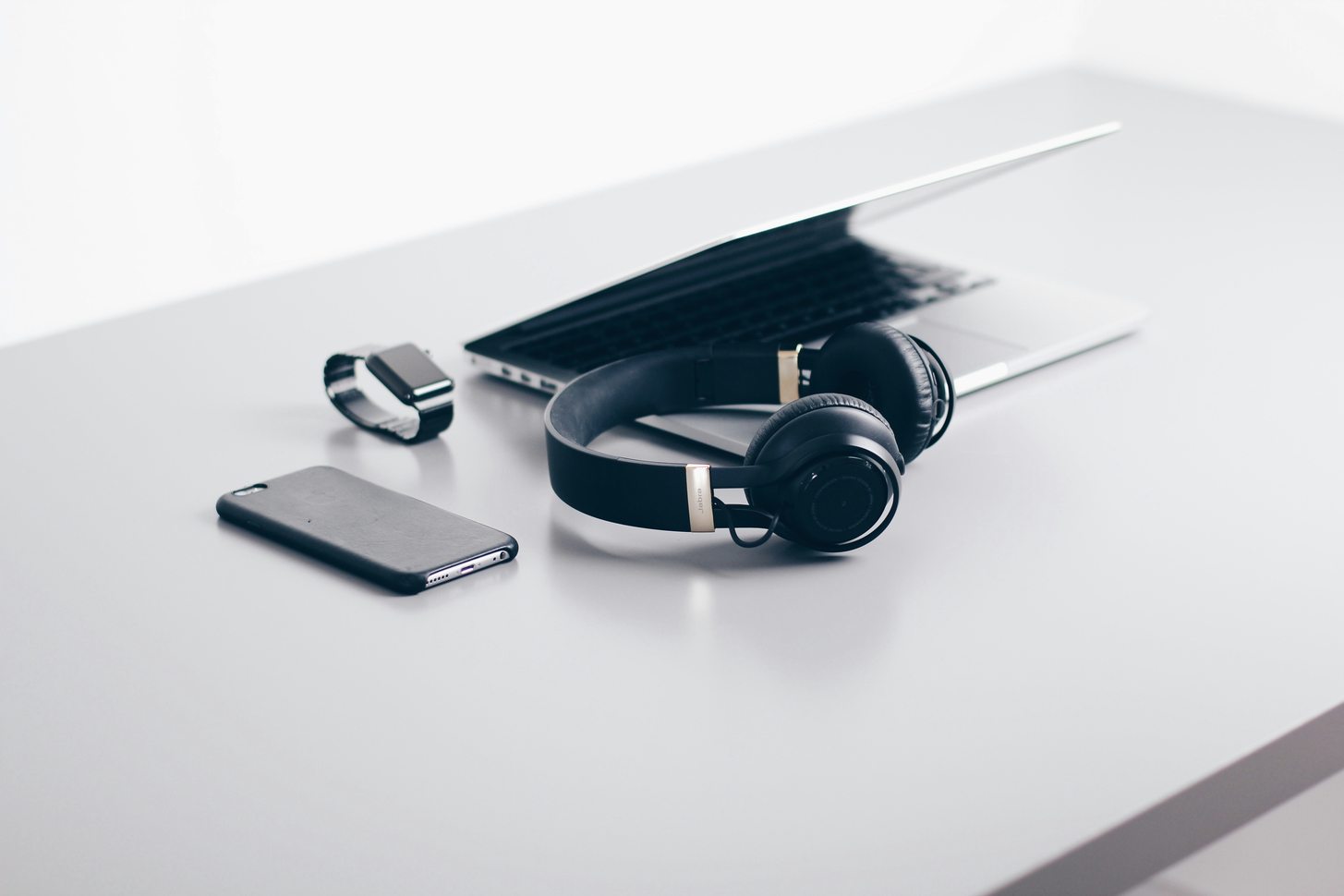 Headphones, a phone and a smartwatch laid out beside a laptop on a pale desk